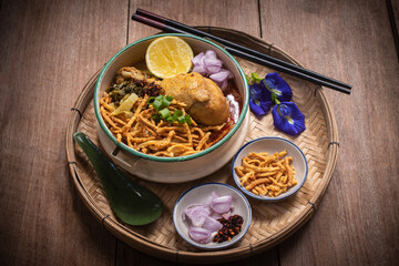 Beautiful still life of traditional Northern Thai Khao Soi chicken curry noodles served in an enamel bowl on a wooden table, with crispy noodles, shallots, lime, chili oil.