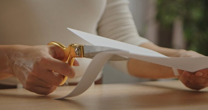 A close-up image of hands using gold scissors to cut a sheet of white paper on a wooden table.