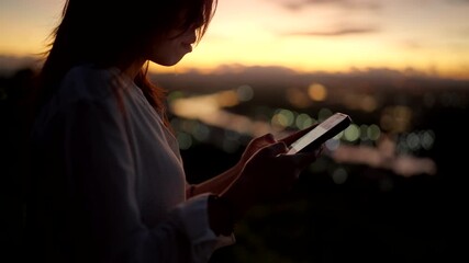 A woman uses her smartphone for social media communication during sunset with city lights in the background. - Powered by Adobe