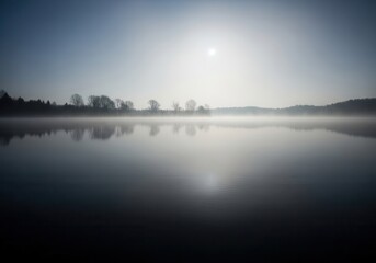 Obraz premium A foggy lake with trees in the distance and the sun in the sky