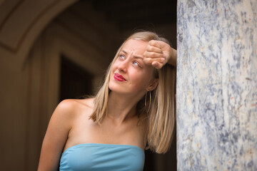 Fototapeta premium Portrait of a young woman, blonde, blue eyes, slim, wearing a light blue dress, innocently looking up at the sky with curiosity, leaning against a marble column. Concept: femininity, innocence, youth.