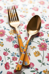 a stylish and premium set of cutlery, consisting of a dinner fork and a table spoon, crossed dynamically against a bright, floral-patterned background. The flatware boasts a stunning gold-tone finish.