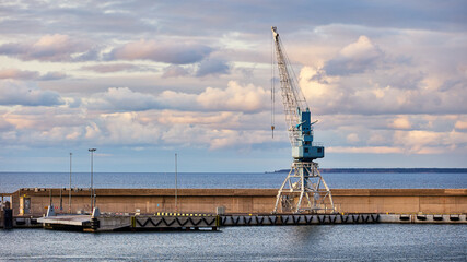 Industrial seaport landscape with a large cargo crane on a pier against a cloudy sky over the Baltic Sea. Calm water, concrete structures, maritime transport and harbor infrastructure.