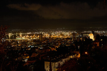 Night panoramic view of Haifa with bright port lights, city skyline, and illuminated Baha'i Gardens overlooking the bay.
