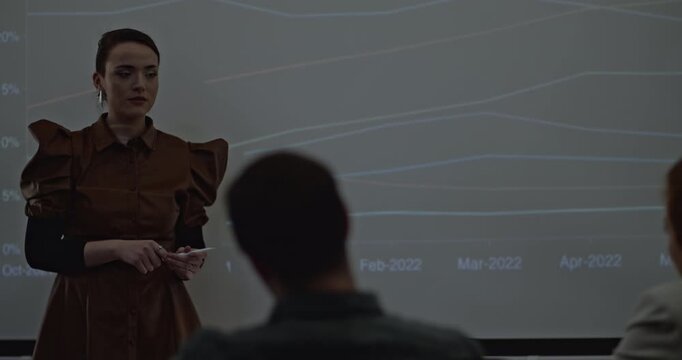 Young business associates analyze line graphs projected on a screen during an office presentation. The presenter holds a stylus, presumably to annotate the data.