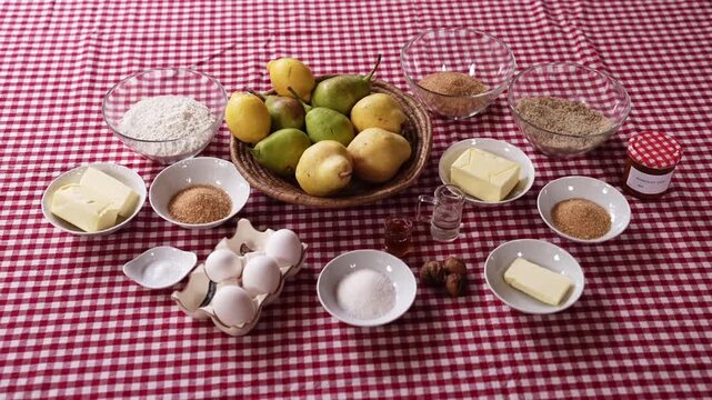 Flat lay of fresh pears, flour, eggs, butter, sugar, nuts, and spices arranged on a red gingham tablecloth, prepared for baking a traditional pear dessert or cake.