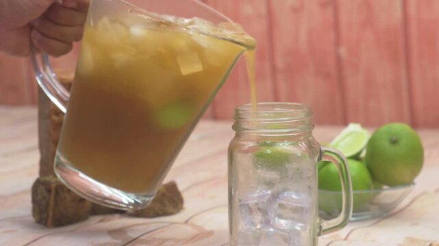 Refreshing close-up hand preparing to pour homemade Papel&oacute;n con Lim&oacute;n into a glass mason jar filled with ice, amidst fresh limes and raw sugar chunks.