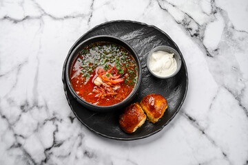 Traditional Ukrainian borscht soup with beetroot, cabbage, pork, fresh herbs, served in black ceramic bowl with sour cream and homemade golden buns on marble background