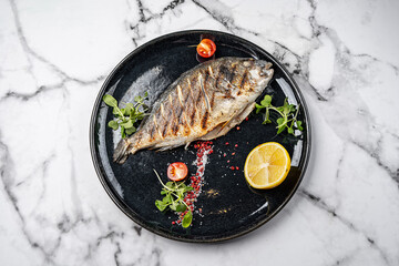 Grilled whole dorado fish served on black plate with lemon half, cherry tomatoes, pink pepper, sea salt and fresh microgreens on marble background