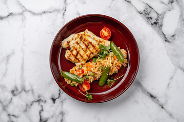 Grilled chicken breast served with vegetable risotto, green peas, cherry tomatoes, and microgreens on a red ceramic plate on marble background