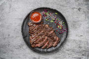 Grilled sliced beef steak served on black plate with spicy red sauce, microgreens, salt and pink pepper on rustic background
