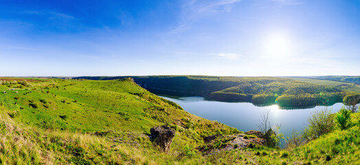 A stunning high-angle panoramic landscape of a wide river canyon near Subich. A lush green, grassy hill slopes down to the calm water, with a dense forest on the opposite bank under a clear blue sky.
