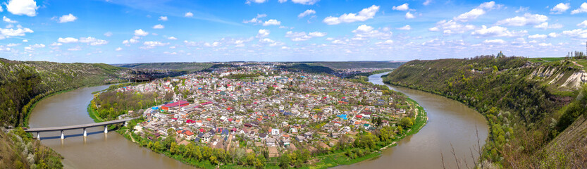 A stunning high-angle panoramic view of Zalishchyky, Ukraine, located in a dramatic meander of the Dniester River. The town is surrounded by steep green hills under a bright blue sky with white clouds