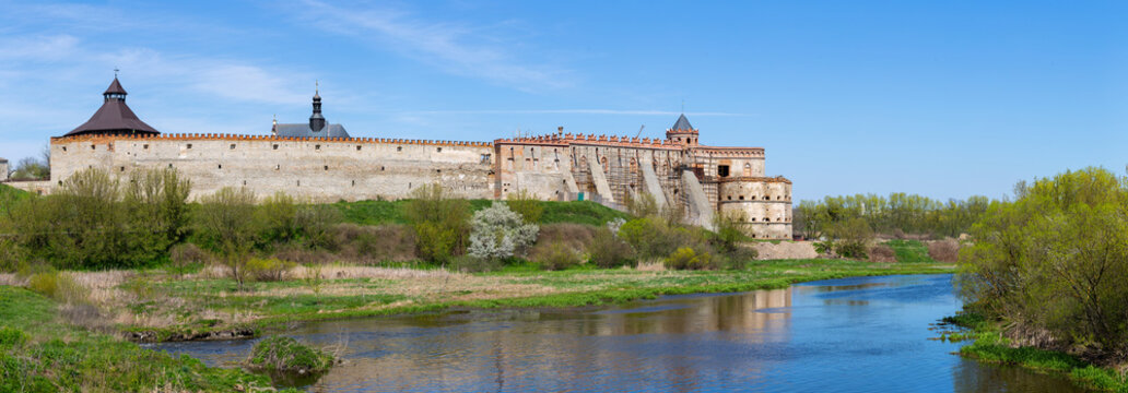 A wide panoramic view of the historic Medzhybizh Castle in Khmelnytskyi Oblast, Ukraine. A large stone fortress with high walls and towers on the bank of the Southern Buh River under a blue sky. - Powered by Adobe