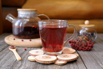 A glass mug of hot herbal tea with berries and fruit stands on an old wooden table against the log...