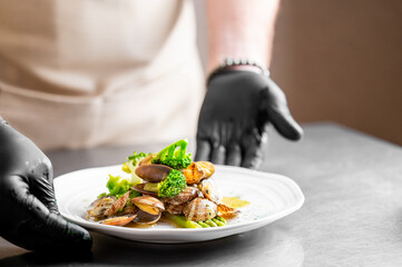 A chef, wearing black gloves, presents a gourmet seafood dish with clams, broccoli, and asparagus on a white plate. The dish is elegantly plated on a metallic surface in a restaurant kitchen setting