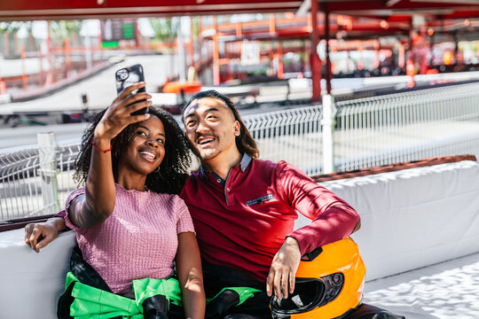 Diverse couple taking a selfie with a smartphone at a go kart track, smiling and enjoying leisure time