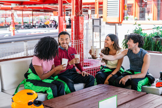 Diverse group of friends wearing racing suits cheering with paper cups, enjoying a break at a go kart track