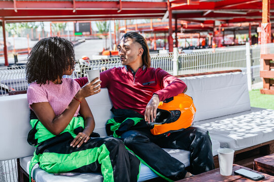Diverse couple drinking coffee and talking, relaxing on a sofa at a go kart track, wearing racing suits before or after racing