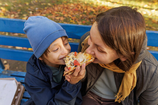 Mother and son playfully sharing pizza slice sittingon park bench in autumn