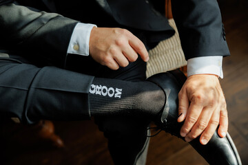 Close-up of groom in elegant black suit adjusting shoes, showing fun socks with “Groom” text. Stylish wedding detail capturing personality and charm of the big day preparation