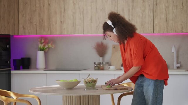 Happy woman in headset listening to music while cutting avocado for fresh salad