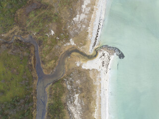 Aerial view of a sinuous stream meeting the pale turquoise sea, where the rugged coastline contrasts with the smooth water, Reine, Nordland, Norway.