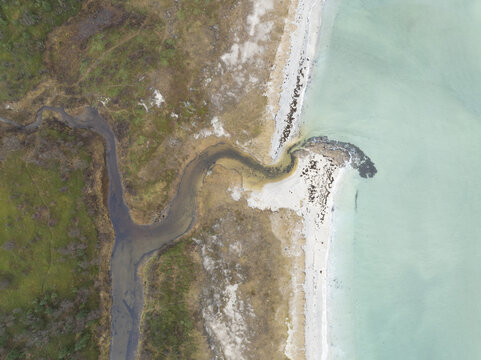 Aerial view of a sinuous stream meeting the pale turquoise sea, where the rugged coastline contrasts with the smooth water, Reine, Nordland, Norway.