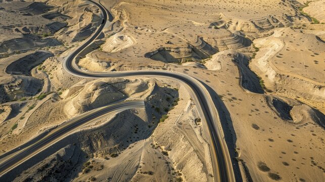 Aerial view of a winding road through a dry, desert landscape. The terrain features rocky hills and sparse vegetation under a clear blue sky.