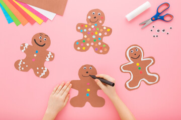 Little child girl hand holding marker and drawing smiling face on brown gingerbread man shapes. Pink table background. Pastel color. Making decoration from application paper. Flat lay. Top down view.