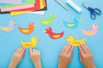 Mother and little child hands creating duck shapes. Scissors, glue stick, pencil and colorful application paper on blue table background. Pastel color. Closeup. Point of view shot. Top down view.
