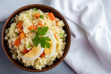 Couscous with vegetables and a piece of egg in a clay bowl on a gray background. Top view. Copy space