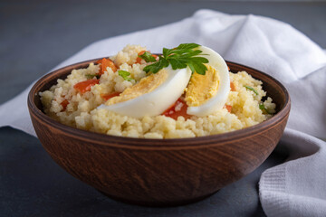 Couscous with vegetables and a piece of egg in a clay bowl on a gray background. 
