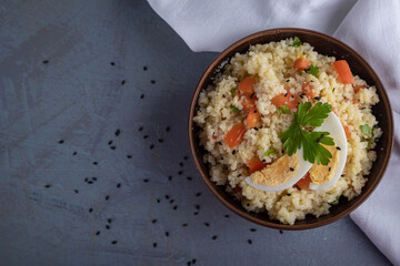 Couscous with vegetables and a piece of egg in a clay bowl on a gray background. Top view. Space for text.