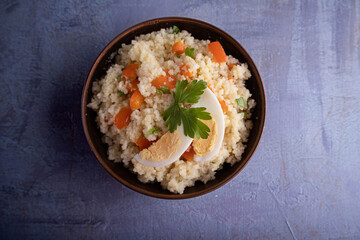 Couscous with vegetables and a piece of egg in a clay bowl on a gray background. Top view. Space for text.