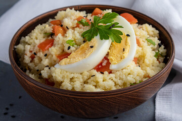 Couscous with vegetables and a piece of egg in a clay bowl on a gray background. 