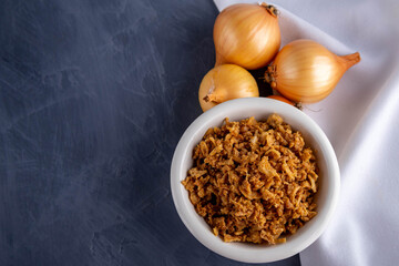 Fried onions in a bowl next to a white towel on a gray background. Top view.