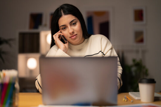 Young Arab female remote employee in casual clothes speaking via smartphone with client while working on project remotely using laptop