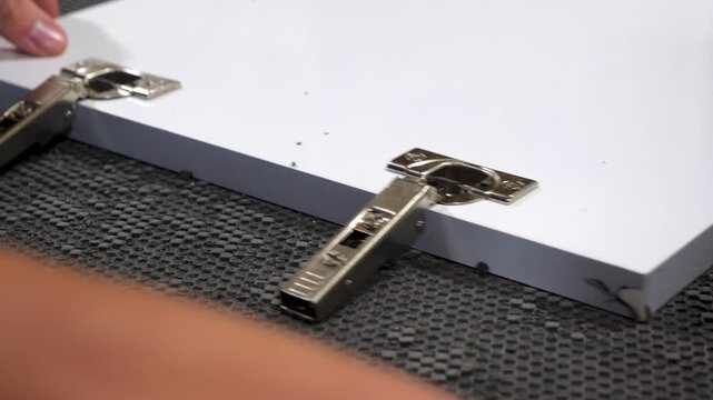 Close-up shot of a hand using a hammer to install a metal hinge on a white wooden panel, set on a textured workbench.