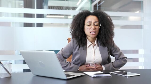 Unwell african american businesswoman suffering from stomach pain sitting at workplace in business office. Unhealthy black woman employee has gastritis, poisoning digestive problems or menstruation