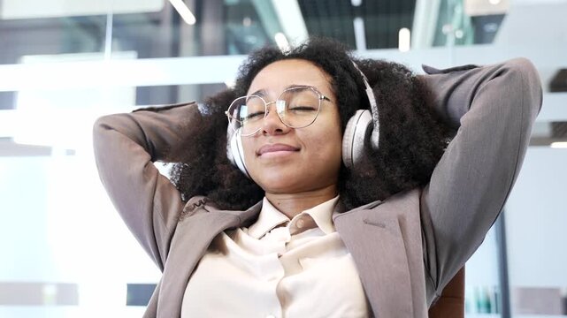 African american businesswoman in headphones listening to relaxing music with eyes closed in office. Woman puts her hands behind her head, relaxes to sounds of relaxation and noise of nature Close up