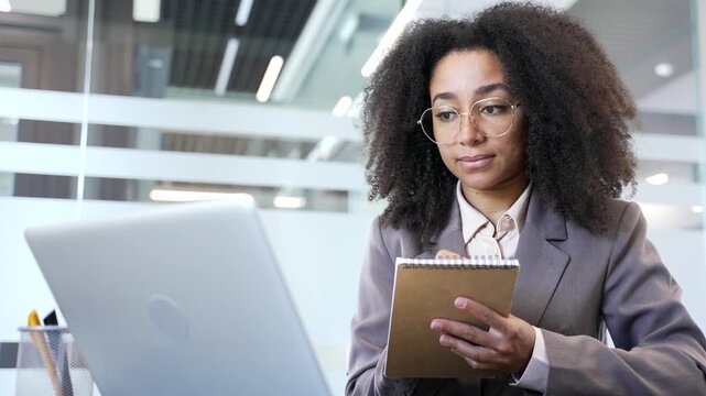 African american businesswoman watching video call online conference taking notes looking at laptop screen while sitting at workplace in office. Black woman listening remote business training Close up