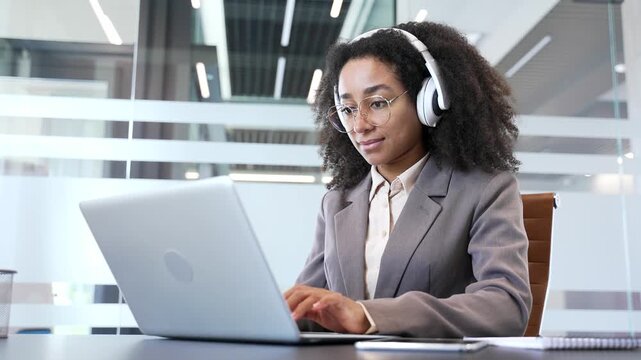 African american businesswoman in wireless headphones typing on laptop sitting at workplace in a modern glass business office. Thoughtful female IT specialist or developer works on project on computer
