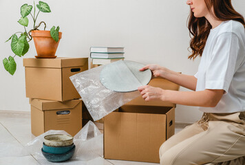 Unrecognized woman wearing white t-shirt sitting on the floor surrounded with carton boxes and packing plates and dishes. Relocating, buying new house. Pack personal stuff from carton boxes. 