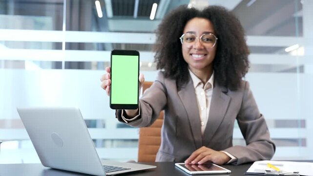 Smiling african american businesswoman showing mobile phone smartphone with green screen sitting at workplace in business office looking at camera. Color key, vertical template layout for advertising