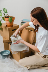 Portrait of woman wearing white t-shirt sitting on the floor surrounded with carton boxes and packing plates and dishes. Relocating, buying new house. Pack personal stuff from carton boxes. 