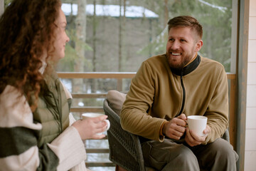 Fototapeta premium Caucasian young adult man smiling and talking while holding coffee cup, sitting outdoors with Caucasian young adult woman, both engaging in relaxed conversation on porch