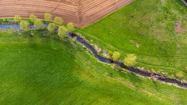 Aerial view of a rural landscape with a small winding stream flowing between green grassy fields and a plowed agricultural area.