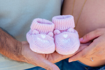 Close up of pregnant couple holding pink baby booties on hands outdoors, concept of maternity, family love, expectation, new life and happiness during pregnancy, symbol of future baby girl arrival.