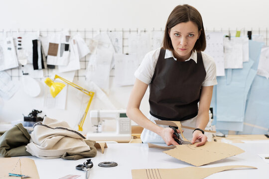 Caucasian female custom tailor cutting out sewing patterns for new clothes in atelier and looking at camera